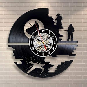May include: Black vinyl record clock featuring Star Wars characters and a Death Star design. The clock has a white face with black numbers and a visible mechanical movement. The clock is mounted on a white brick wall.