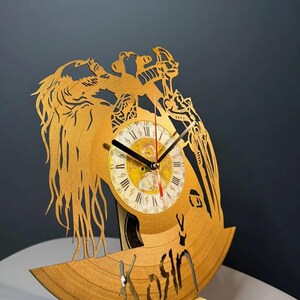 May include: A wooden clock made from a repurposed vinyl record. The clock features a laser-cut design of a figure with long hair and a bow and arrow. The clock face has Roman numerals and black clock hands. The word "Kosin" is cut into the record.