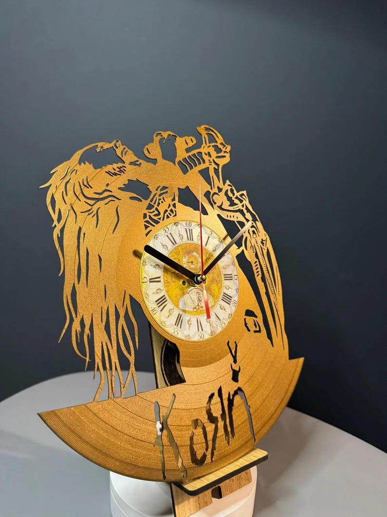 May include: A wooden clock made from a repurposed vinyl record. The clock features a laser-cut design of a figure with long hair and a bow and arrow. The clock face has Roman numerals and black clock hands. The word "Kosin" is cut into the record.