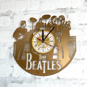 Può includere: Un orologio da parete color oro con una silhouette dei membri della band The Beatles che suonano strumenti. Il quadrante dell'orologio è bianco con numeri romani e lancette nere. Il nome della band, "THE BEATLES", è ritagliato in basso.