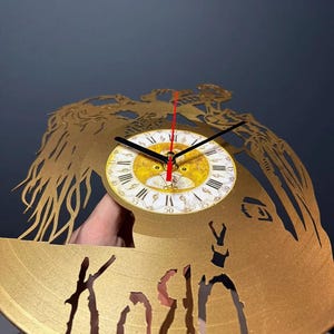 May include: Gold-colored wall clock with a cut-out design featuring a face and the band name "Korn". The clock face is white with Roman numerals and black hands. The second hand is red.