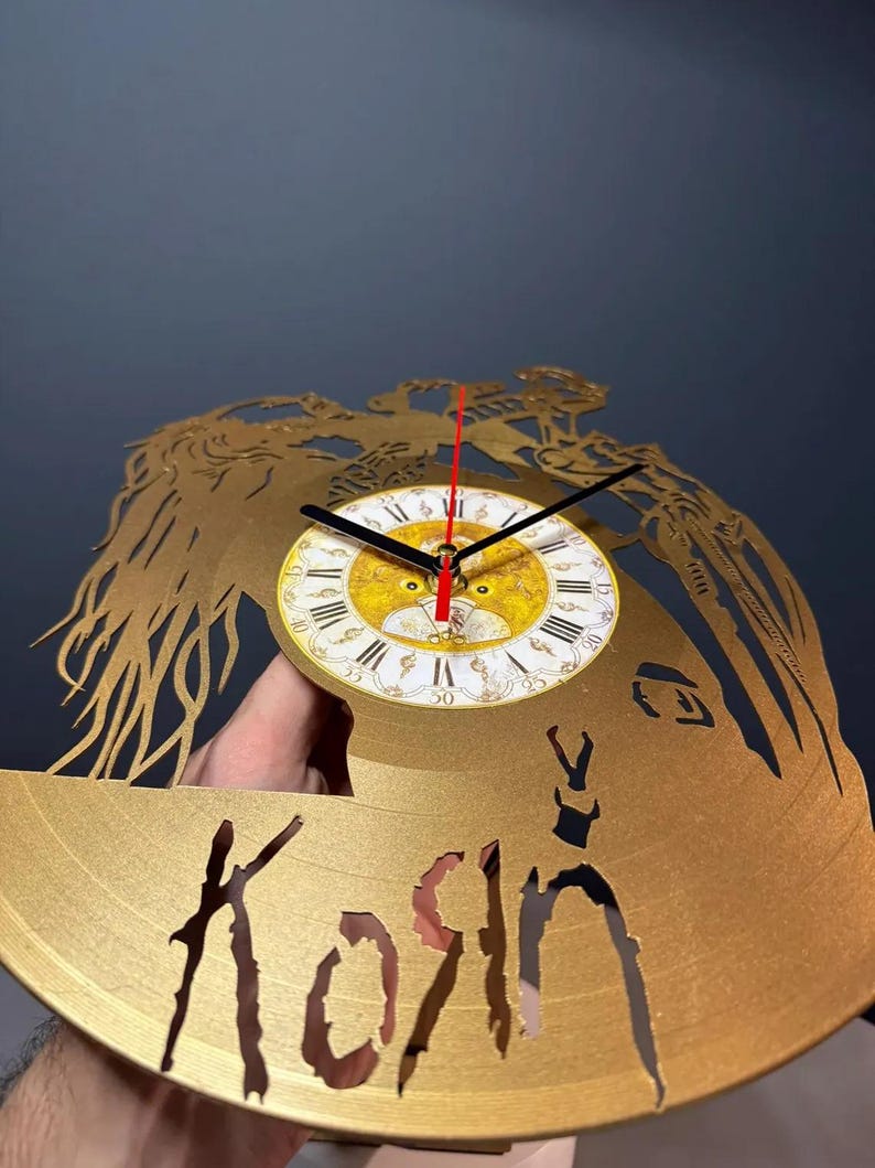 May include: Gold-colored wall clock with a cut-out design featuring a face and the band name "Korn". The clock face is white with Roman numerals and black hands. The second hand is red.