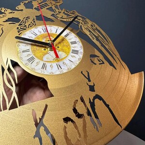 May include: Gold-colored wall clock with a Roman numeral clock face. The clock hands are black, and the second hand is red. The clock has a unique design with cut-out shapes and the word "Korn" visible. The clock is approximately 12 inches in diameter.