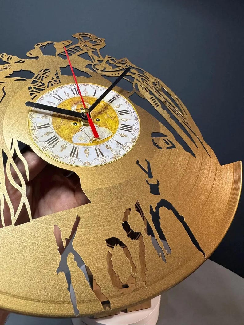 May include: Gold-colored wall clock with a Roman numeral clock face. The clock hands are black, and the second hand is red. The clock has a unique design with cut-out shapes and the word "Korn" visible. The clock is approximately 12 inches in diameter.
