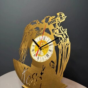 May include: A gold-colored, laser-cut clock made from a repurposed vinyl record. The clock features a detailed design with a central clock face, Roman numerals, and the word "Koshin" cut into the record. The clock is supported by a wooden stand.