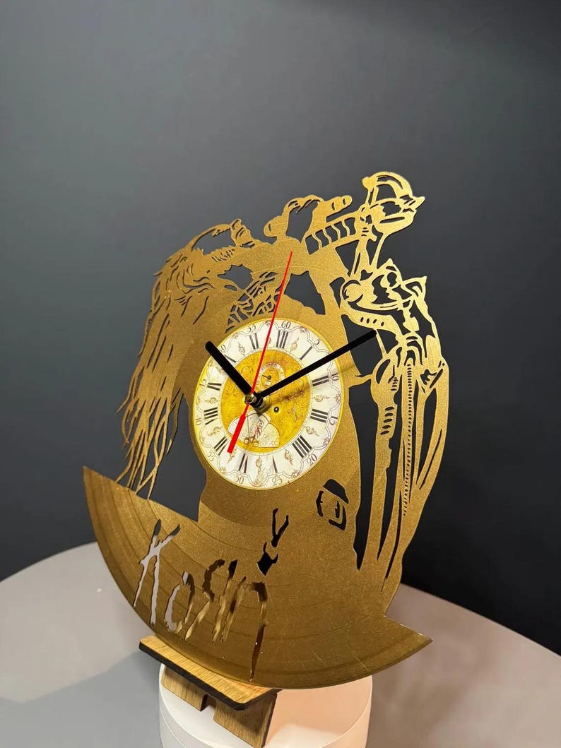 May include: A gold-colored, laser-cut clock made from a repurposed vinyl record. The clock features a detailed design with a central clock face, Roman numerals, and the word "Koshin" cut into the record. The clock is supported by a wooden stand.