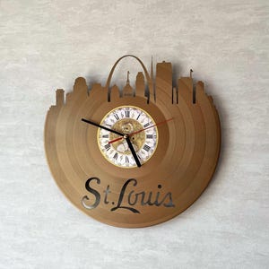 Op de afbeelding: Een bruine houten klok in de vorm van een vinylplaat, met de skyline van St. Louis. De klok heeft Romeinse cijfers en zwarte wijzers. De naam van de stad "St. Louis" is eronder uitgesneden.