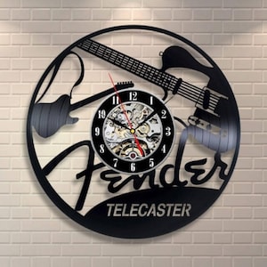 May include: Black wall clock with a guitar silhouette design, the Fender logo, and the word "Telecaster". The clock face has a visible mechanical movement and red hands. The clock is mounted on a white brick wall.