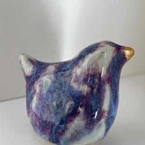 May include: A ceramic bird ornament with a rounded body and a small tail. The bird is glazed with a marbled pattern of blue, purple, and white. It has a gold-coloured beak.