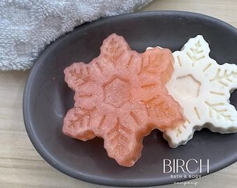 Puede incluir: Dos jabones para baño y cuerpo con forma de copo de nieve, uno es de color rosa claro y el otro es blanco. Los jabones están en un plato ovalado gris oscuro. El texto "BIRCH BATH & BODY company" está impreso en el plato.