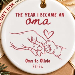 May include: A round white ceramic ornament featuring a red line drawing of an adult hand holding a baby hand with a heart. The text reads: THE YEAR I BECAME AN oma, Oma to Olivia, 2026. A gold ribbon is attached to the top for hanging purposes.
