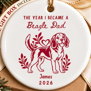 May include: A round white ceramic ornament featuring a red graphic of a beagle dog, a heart, and leafy sprigs. The text reads THE YEAR I BECAME A Beagle Dad, James, and 2026. A gold cord is attached to the top, and a beige ribbon is tied behind it.
