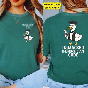 May include: A teal t-shirt with a cartoon duck wearing a graduation cap and holding a scroll. The front of the shirt says "Mortician Emily Est. 2021". The back of the shirt reads "I Quacked the Mortician Code".