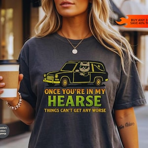 May include: A charcoal gray t-shirt featuring a graphic of a cat wearing sunglasses driving a black hearse. The text reads: ONCE YOU'RE IN MY HEARSE THINGS CAN'T GET ANY WORSE. The shirt has a crew neckline and short sleeves, with a Comfort Colors Pepper label.