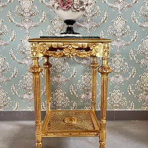 May include: A gold-colored, ornate side table with a marble top and a lower shelf. The table supports a white and black decorative vase filled with pink and white floral accents. The background features a patterned wallpaper.
