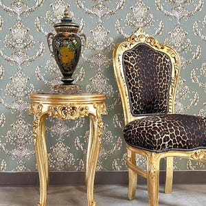 May include: A gold-colored side table with ornate carvings supports a decorative vase. A matching gold chair with leopard print upholstery sits nearby. The background features a patterned wallpaper in shades of green and gold.