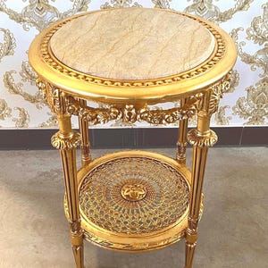 May include: A small, ornate gold side table with a round, beige marble top. The table features intricate carvings, a lower shelf with a decorative lattice design, and stands on four legs. A classic piece of furniture.