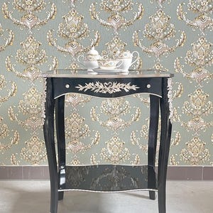 Peut inclure: Une table d'appoint noire avec des accents argentés et deux étagères. La table est décorée d'un motif floral argenté et a un plateau en verre. Il y a trois tasses à thé en porcelaine et soucoupes sur l'étagère supérieure.