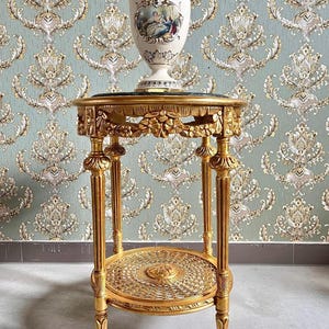 Może przedstawiać: Okrągły, dwupoziomowy stolik pomocniczy pomalowany na złoto z blatem z zielonego marmuru. Stolik ma misterne rzeźby i pleciony dolny półkę.