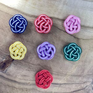 May include: Seven colorful, decorative, woven knots in the shape of flowers. The knots are in shades of red, yellow, green, purple, pink, and blue.