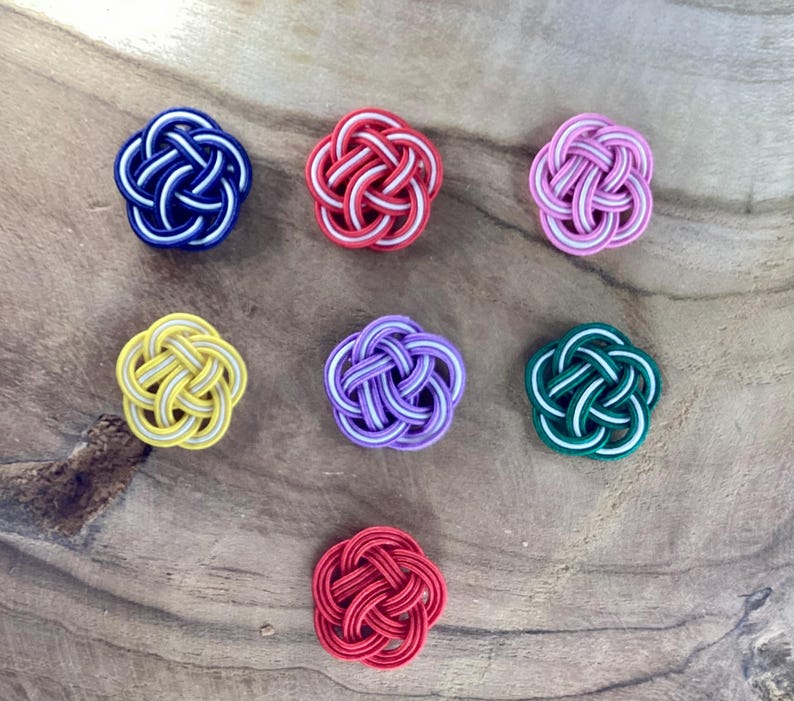 May include: Seven colorful, decorative, woven knots in the shape of flowers. The knots are in shades of red, yellow, green, purple, pink, and blue.