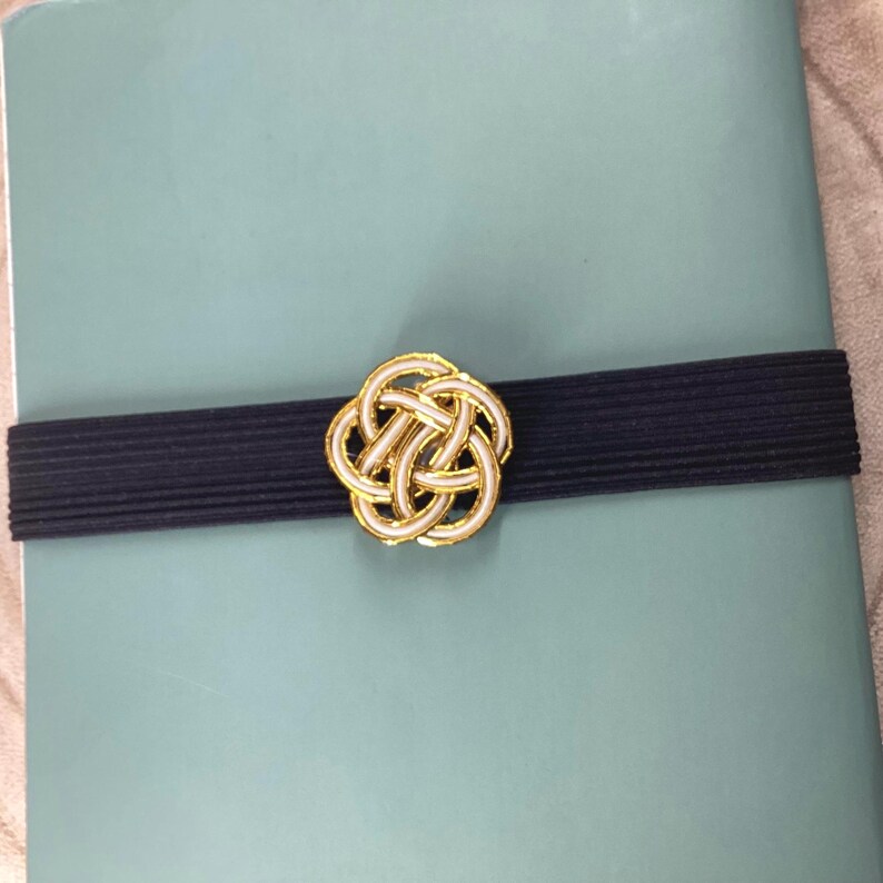 May include: A gold and white decorative knot design on a dark blue elastic band. The band is on a light blue surface.
