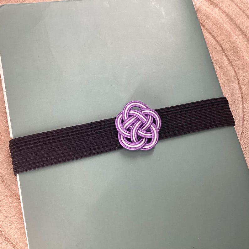 May include: A purple and white decorative knot tied with a black elastic band. The knot is a traditional Japanese design.