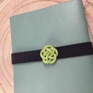 May include: A green and white decorative knot tied with a black elastic band. The knot is a traditional Japanese design.