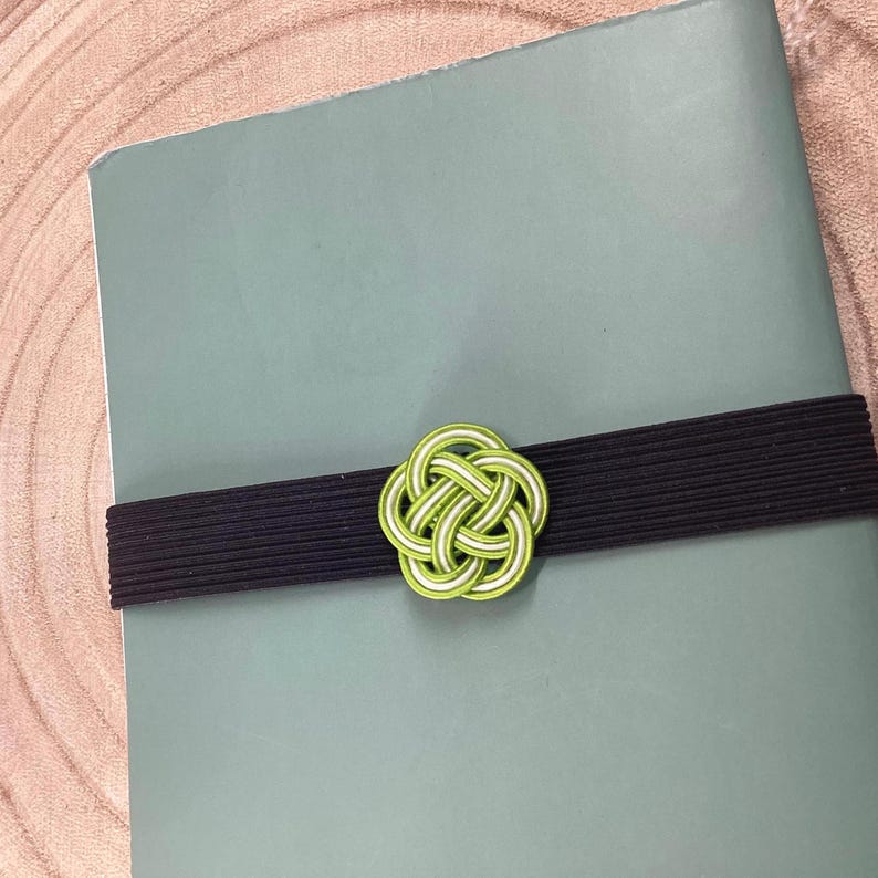 May include: A green and white decorative knot tied with a black elastic band. The knot is a traditional Japanese design.