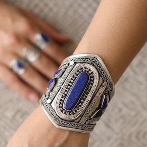 May include: A silver cuff bracelet with intricate detailing and inlaid rectangular blue stones. The bracelet is worn on a wrist, with rings featuring similar blue stones visible in the background. The jewelry has an antique or vintage aesthetic.