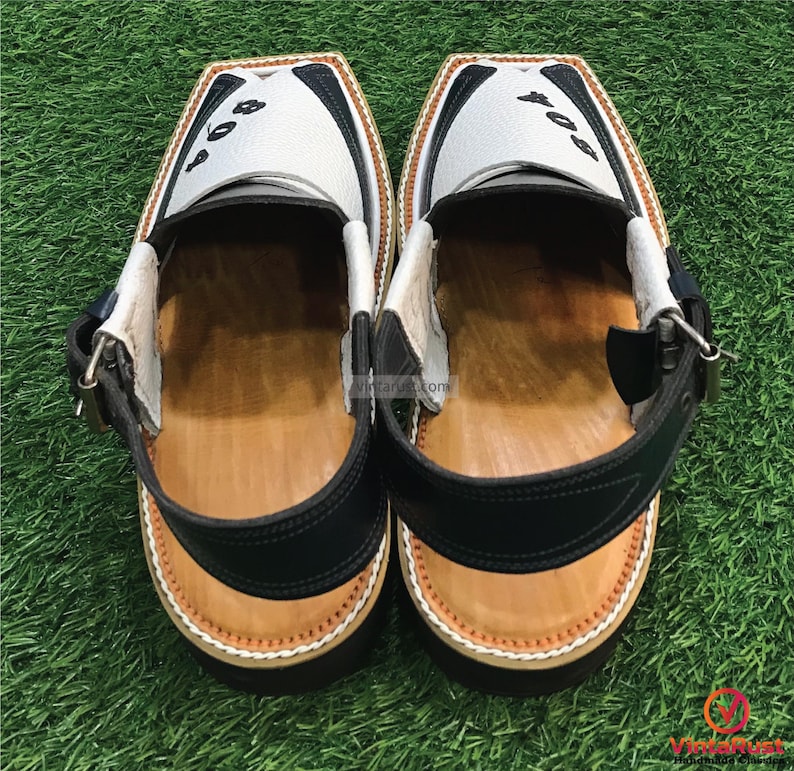 May include: A pair of white leather sandals with black accents and a brown leather insole. The sandals have a strap with a buckle and the number "804" is written on the top of each sandal.