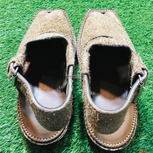 May include: A pair of brown suede sandals with a strap and buckle. The sandals have a brown leather sole and are on a green grassy surface.