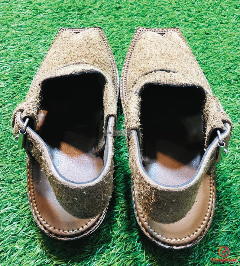 May include: A pair of brown suede sandals with a strap and buckle. The sandals have a brown leather sole and are on a green grassy surface.