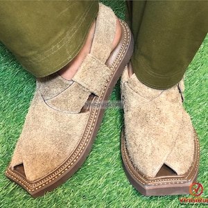 May include: A pair of brown suede sandals with a pointed toe and decorative stitching around the edge of the sole. The sandals are being worn on a person's feet, with their legs crossed.