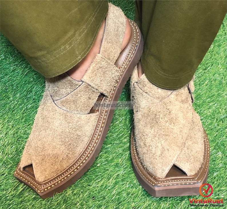 May include: A pair of brown suede sandals with a pointed toe and decorative stitching around the edge of the sole. The sandals are being worn on a person's feet, with their legs crossed.