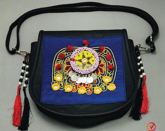 May include: A black leather shoulder bag with a blue fabric flap featuring colorful embroidery, silver coins, and mirror accents. The bag has a long black strap and red and black tassels.
