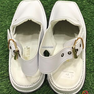 May include: White leather sandals with a gold buckle and a white strap. The sandals have a white sole and are labeled "Bigger Khan" with the size "42" on the bottom.