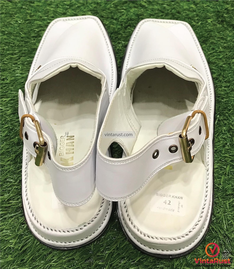May include: White leather sandals with a gold buckle and a white strap. The sandals have a white sole and are labeled "Bigger Khan" with the size "42" on the bottom.
