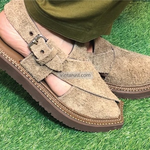 May include: A pair of brown leather sandals with a buckle strap and a thick sole. The sandals are made of suede and have a textured finish. The sandals are on a green grassy surface.