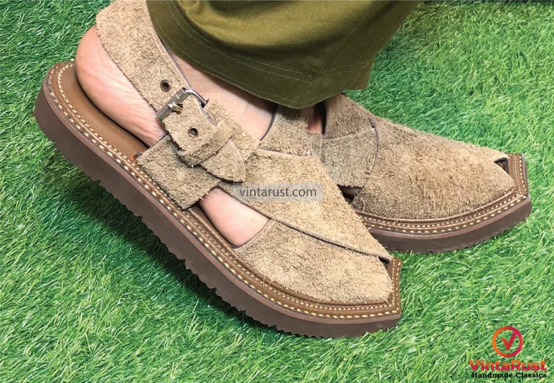 May include: A pair of brown leather sandals with a buckle strap and a thick sole. The sandals are made of suede and have a textured finish. The sandals are on a green grassy surface.