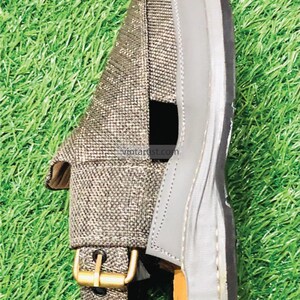 May include: A pair of gray leather sandals with a woven fabric strap and a buckle closure. The sandals are on a green grassy surface.