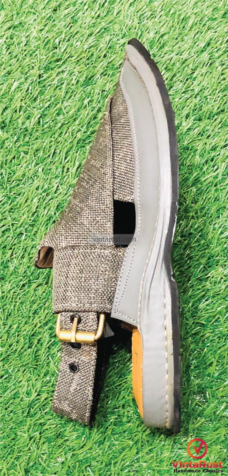 May include: A pair of gray leather sandals with a woven fabric strap and a buckle closure. The sandals are on a green grassy surface.