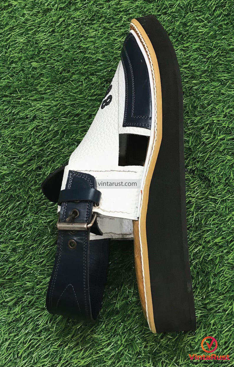 May include: White leather sandals with a blue strap and buckle. The sandals have a thick black sole with a brown edge.
