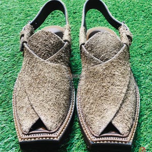 May include: A pair of brown suede sandals with a pointed toe and a strap across the top of the foot. The sandals have a brown leather sole and decorative stitching around the edge.