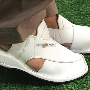May include: White leather sandals with a closed toe and an open heel. The sandals have a thick white sole and a decorative gold buckle.