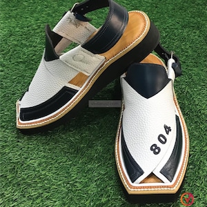 May include: White and navy blue leather sandals with a brown sole. The sandals have a strap across the top and a buckle closure. The sandals have the number "804" printed on the side.