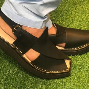May include: Black leather sandals with a closed toe and an open heel. The sandals have a thick sole and a strap that goes around the ankle.
