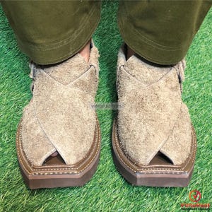 May include: A pair of tan suede sandals with a distinctive design. The sandals have a closed toe and a strap that goes over the top of the foot. The sandals are sitting on a green grassy surface.
