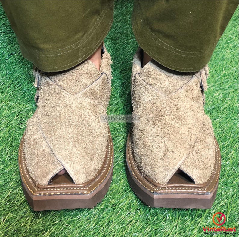 May include: A pair of tan suede sandals with a distinctive design. The sandals have a closed toe and a strap that goes over the top of the foot. The sandals are sitting on a green grassy surface.