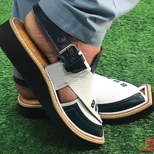 May include: White and blue leather sandals with a black strap and buckle. The sandals have a thick black sole and a white stitched trim.